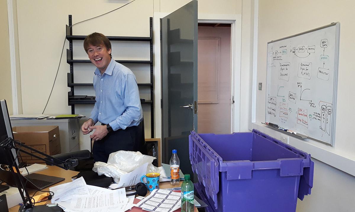 Laughing man in partly empty office, packing up items into purple box.