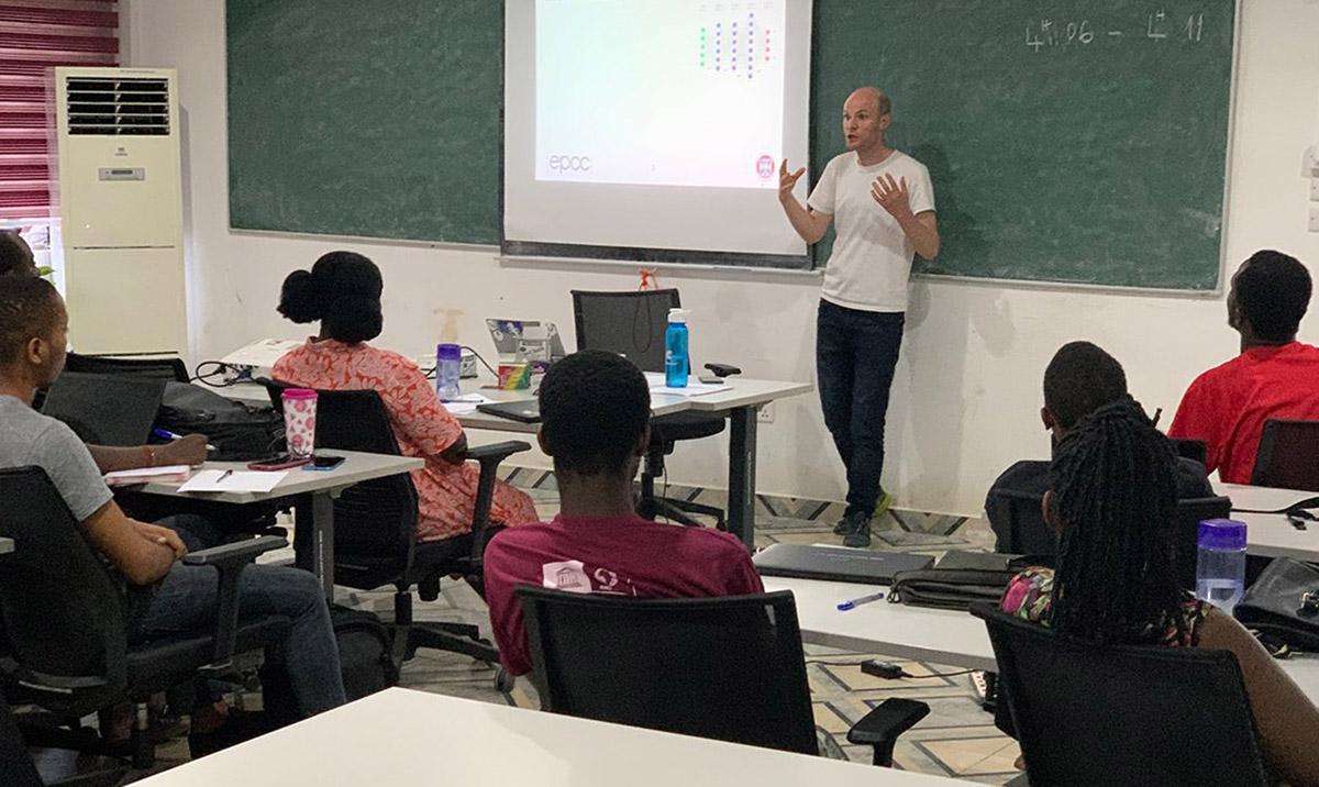 Classroom image showing lecturer standing in front of board, talking to seated students.