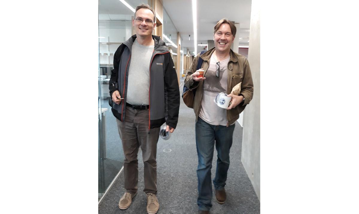 Two smiling men walking down an office corridor.