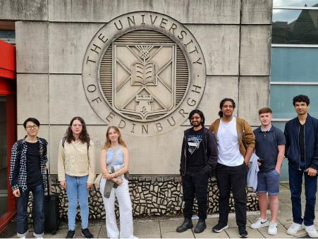7 Masters students standing beside a University logo carved into a sandstone building.