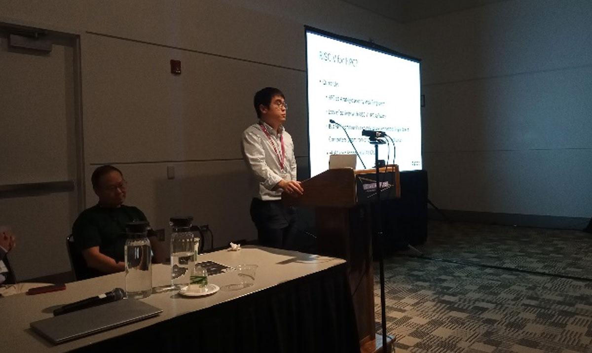 Photo shows man giving presentation beside screen, with another man seated at a desk nearby.