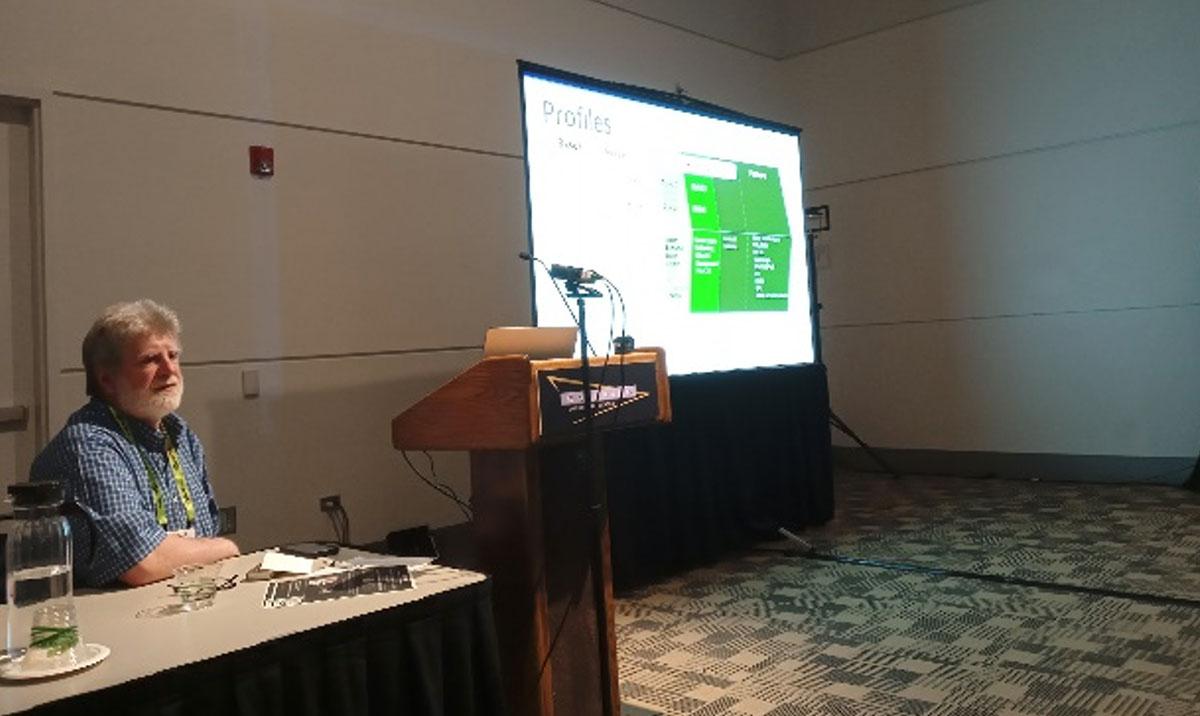 Photo shows seated man giving presentation beside large screen.