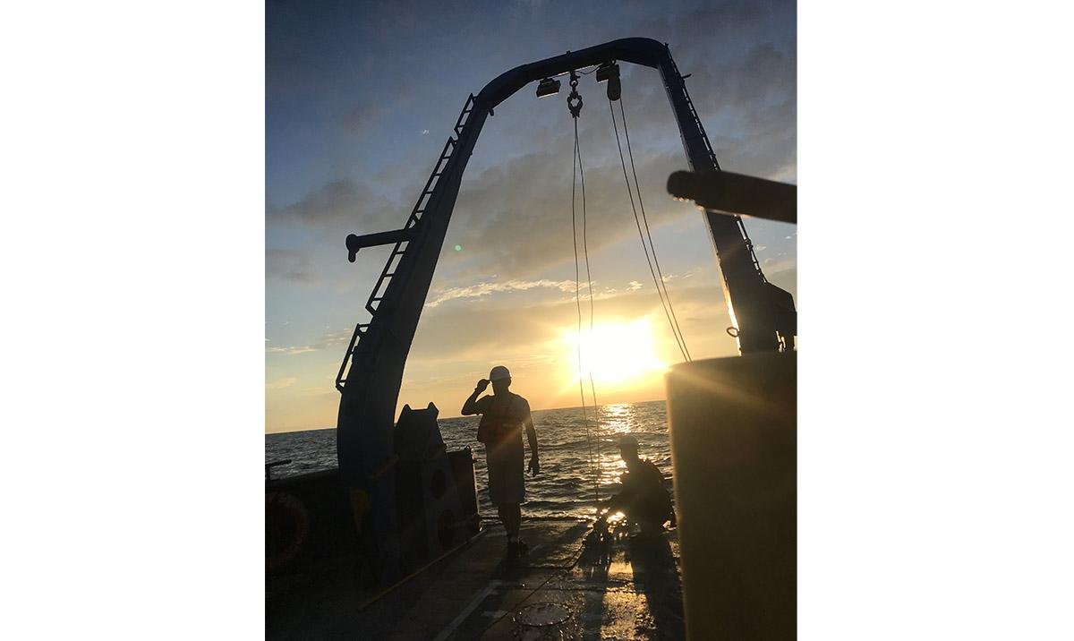 Photo showing 2 men working on ship silhouetted against sunset.