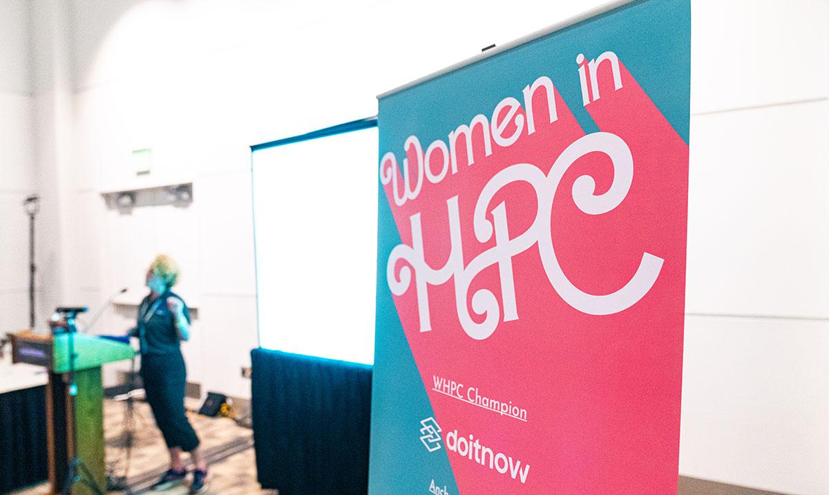 Woman speaking at podium beside "Women in HPC" banner