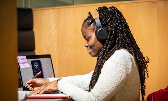 Woman wearing headphones taking notes beside laptop.