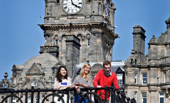 Photograph of two young women and one man leaning against railings. Backdrop is old clock tower and city buildings on a sunny day.